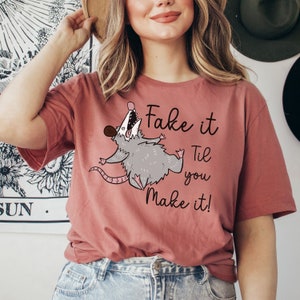 Puede incluir: Una camiseta rosa con un opossum de dibujos animados y el texto "Fake it Til you Make it!"