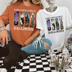 May include: A rust-colored t-shirt and a white t-shirt, both featuring a graphic print with four vertical panels showing a cauldron, a black cat, a haunted house, and a mummy, with the text EASILY DISTRACTED BY HALLOWEEN printed in black.