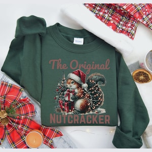 May include: Dark green sweatshirt with a festive graphic. The graphic features a squirrel wearing a Santa hat, holding a gift, and surrounded by Christmas lights and a small tree. The text "The Original Nutcracker" is above and below the squirrel.
