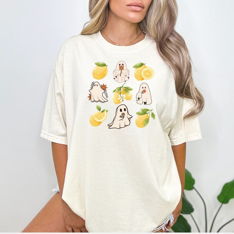 May include: A cream-colored t-shirt with a graphic design of ghosts holding lemons and lemon branches. The ghosts are white and the lemons are yellow.