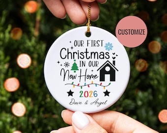 May include: A round white ceramic Christmas ornament featuring the black text Our First Christmas in Our New Home, a green pine tree, a black house icon, a string of colorful lights, the year 2026 in multi-colored digits, and the names Dave & Angel.