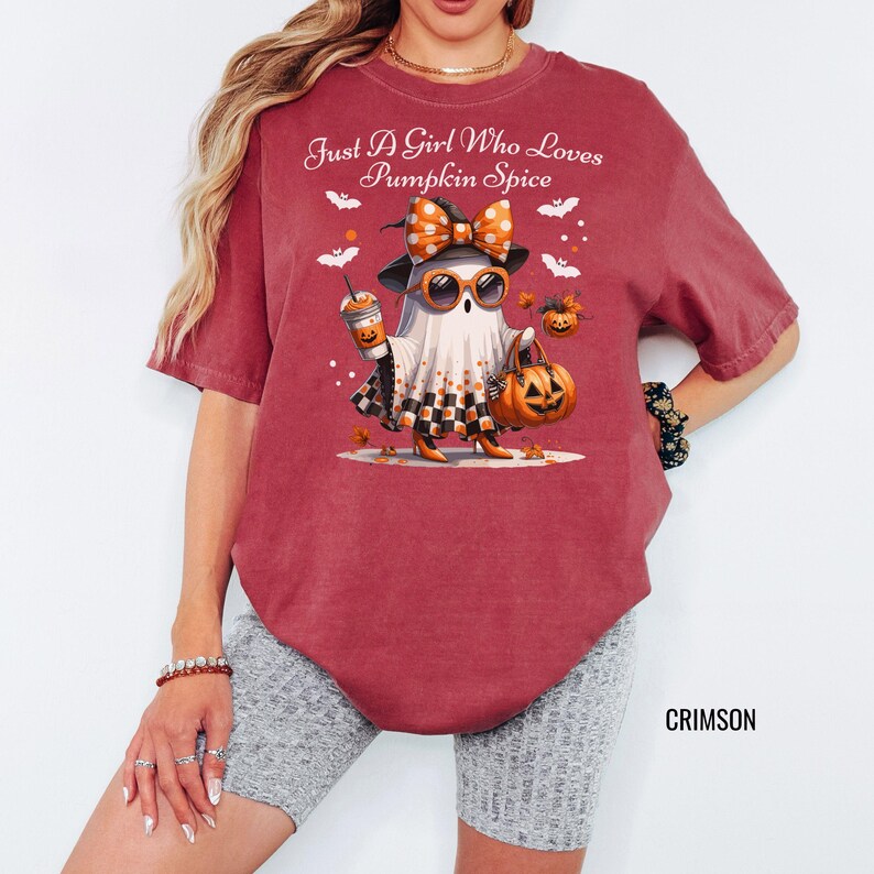 May include: Crimson-colored t-shirt featuring a Halloween-themed graphic. The graphic depicts a ghost character wearing sunglasses, a bow, and holding a pumpkin spice latte and a jack-o'-lantern bag. The text "Just A Girl Who Loves Pumpkin Spice" is above the graphic.