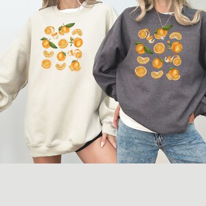 May include: Two sweatshirts with a graphic print of orange slices and leaves. One sweatshirt is cream colored and the other is dark gray.