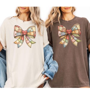 May include: Two t-shirts, one cream and one brown, each featuring a large bow design. The bow is made of patchwork squares in pastel colors, with a red heart in the center that reads "LOVE". The shirts have short sleeves and a relaxed fit.