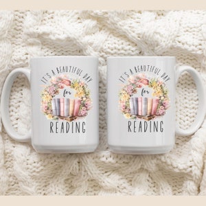 May include: Two white ceramic mugs with a floral wreath design and the text "It's a beautiful day for reading." The design includes a stack of books in pastel colors. The mugs have large handles.