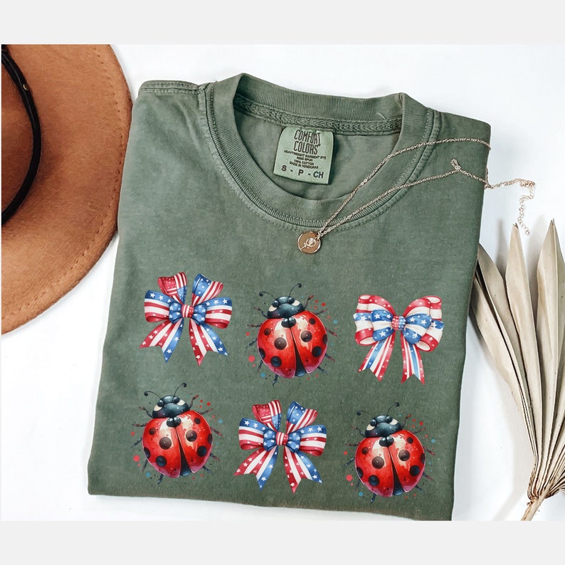 以下が含まれることがあります： 赤と青のリボンとテントウムシのデザインが施された緑色のTシャツ。Tシャツにはタグに「COMBAT COLORS」の文字があります。