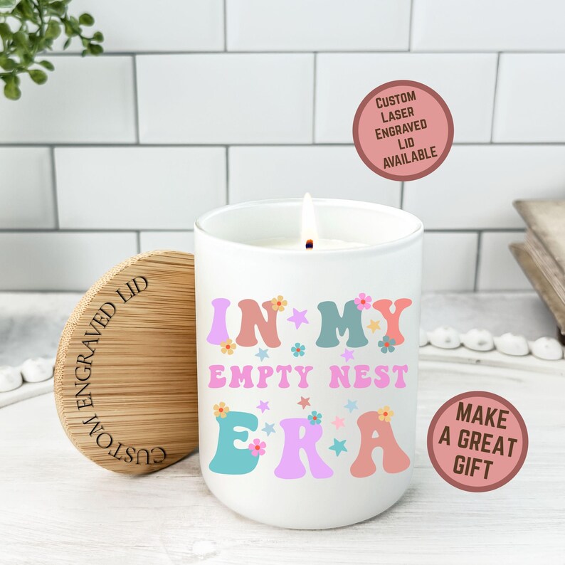 Op de afbeelding: Een witte kaars met de tekst "IN MY EMPTY NEST ERA" in kleurrijke letters en bloemaccenten. De kaars heeft een houten deksel met de woorden "CUSTOM ENGRAVED LID" en een roze sticker met de tekst "MAKE A GREAT GIFT".