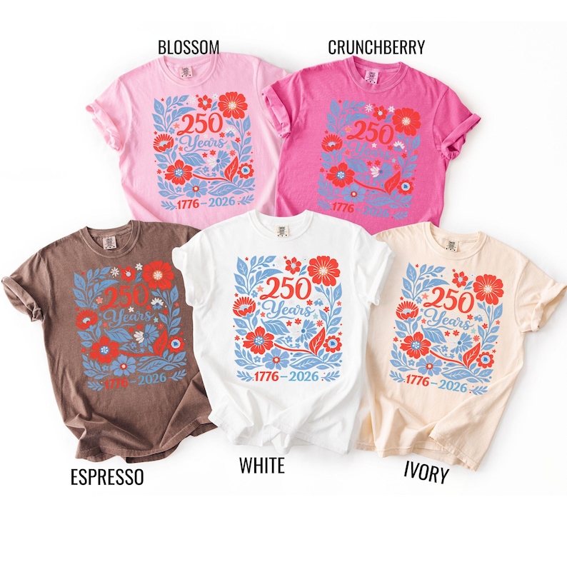 May include: Five t-shirts in various colors: blossom, crunchberry, espresso, white, and ivory. Each shirt features a floral design with the text "250 Years 1776-2026" in red and blue. The shirts are short-sleeved and have a casual style.
