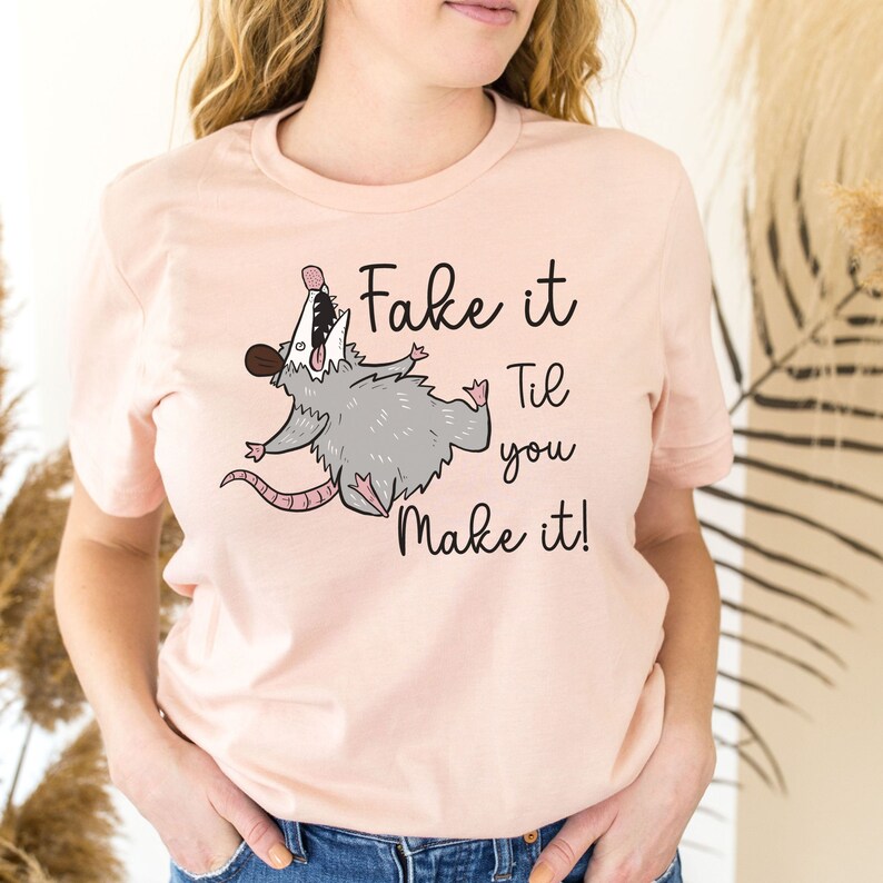 Puede incluir: Una camiseta rosa con un opossum de dibujos animados y el texto "Fake it Til you Make it!"