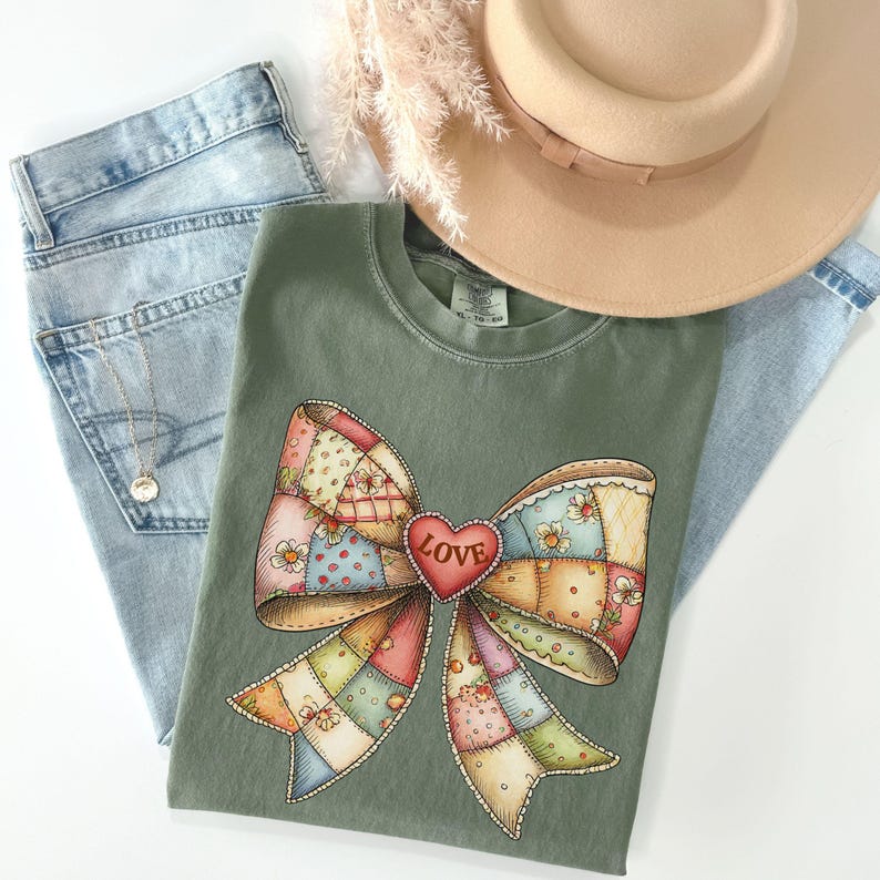 May include: An olive green t-shirt featuring a colorful patchwork bow design with a red heart that says "LOVE". The shirt is paired with light blue jeans, a beige hat, and a delicate necklace. The image showcases a casual, feminine aesthetic.