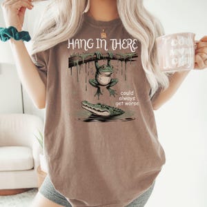 May include: Brown t-shirt with a frog hanging from a branch above an alligator in water. The text "Hang in There" is above the frog, and "could always get worse" is below the alligator. The shirt is a neutral color.