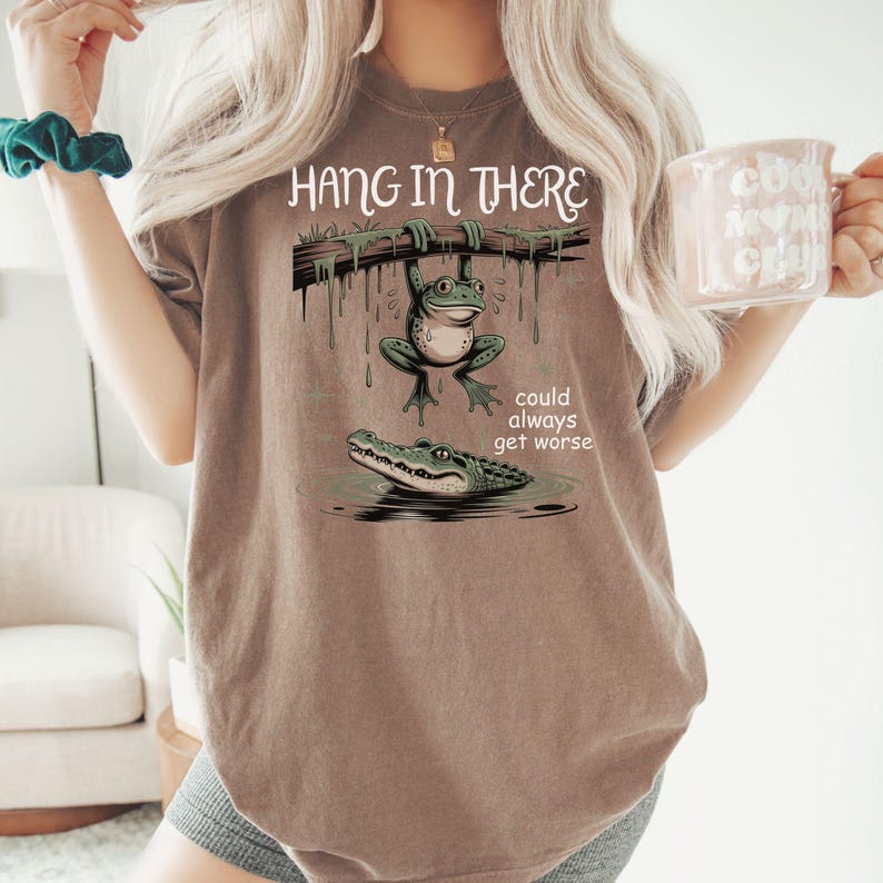 May include: Brown t-shirt with a frog hanging from a branch above an alligator in water. The text "Hang in There" is above the frog, and "could always get worse" is below the alligator. The shirt is a neutral color.