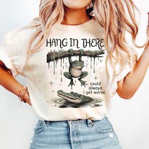 May include: Cream-colored t-shirt featuring a cartoon frog hanging from a branch above an alligator in water. The text "HANG IN THERE" is above the frog, and "could always get worse" is below. The design is in shades of green and brown.