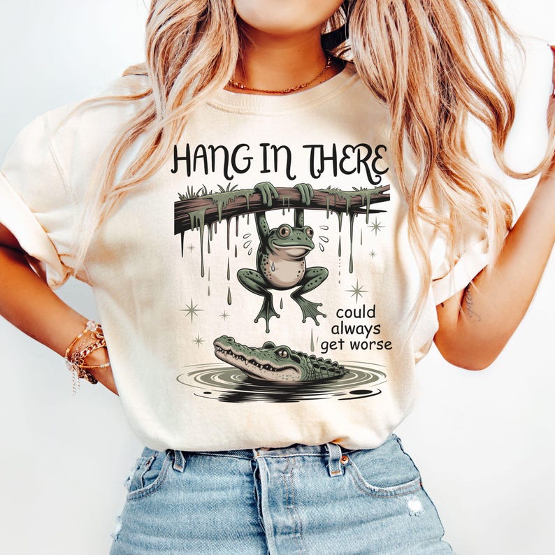 May include: Cream-colored t-shirt featuring a cartoon frog hanging from a branch above an alligator in water. The text "HANG IN THERE" is above the frog, and "could always get worse" is below. The design is in shades of green and brown.