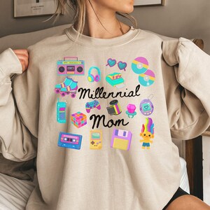 May include: Beige sweatshirt with colorful 90s-themed graphics. Includes a boombox, roller skates, headphones, a cassette tape, a floppy disk, and the text "Millennial Mom".