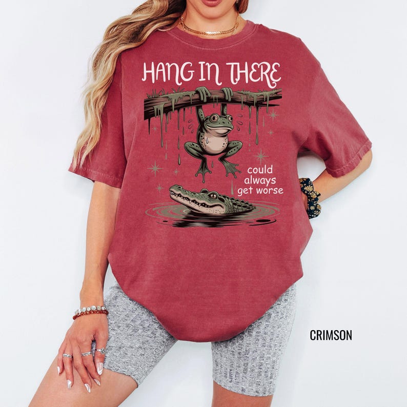 May include: Crimson-colored t-shirt with the phrase "Hang in There" above a cartoon frog hanging from a branch, with an alligator below. The text "could always get worse" is also included. The shirt is a relaxed fit.