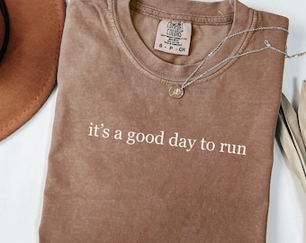 May include: A brown Comfort Colors t-shirt with the phrase "it's a good day to run" in white text. A silver necklace with a small pendant rests on the shirt. The shirt is folded and has a vintage look.