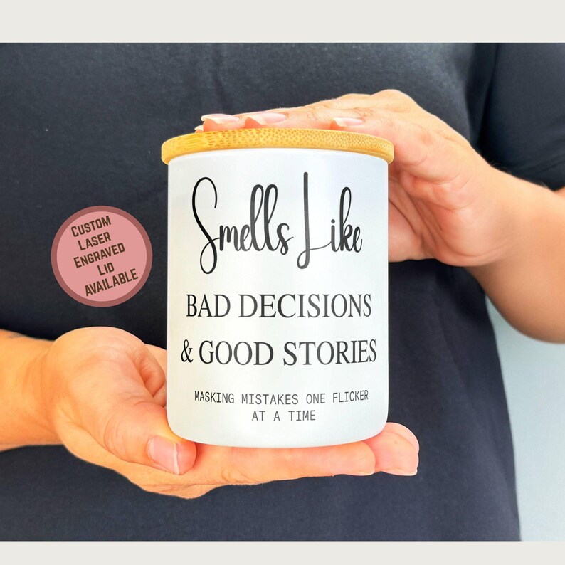 May include: A white candle jar with a bamboo lid is held in the hands. The jar has the text "Smells Like BAD DECISIONS & GOOD STORIES" and "MASKING MISTAKES ONE FLICKER AT A TIME". A pink circle with text "CUSTOM LASER ENGRAVED LID AVAILABLE" is in the image.