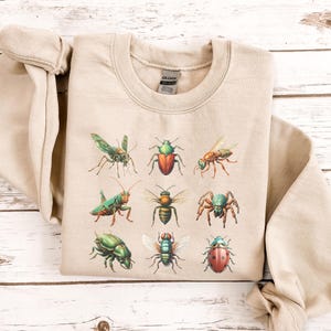 May include: A beige sweatshirt with a graphic print of various insects, including a green grasshopper, a red and black ladybug, a green and black beetle, a green and black spider, a green and black fly, a green and black bee, a green and black butterfly, and a green and black ant.