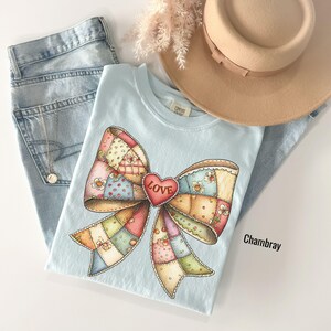 May include: A light blue t-shirt with a large, colorful bow design. The bow is made of patchwork squares and features a red heart with the word "LOVE." The shirt is paired with light wash jeans and a tan hat.