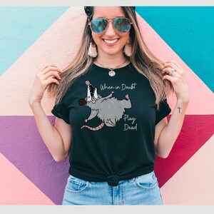 Pu&ograve; includere: Maglietta nera con un opossum dei cartoni animati e il testo "When in Doubt Play Dead".