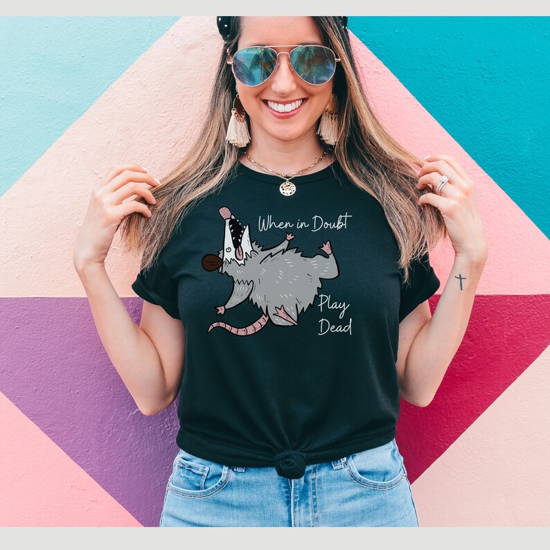 Pu&ograve; includere: Maglietta nera con un opossum dei cartoni animati e il testo "When in Doubt Play Dead".