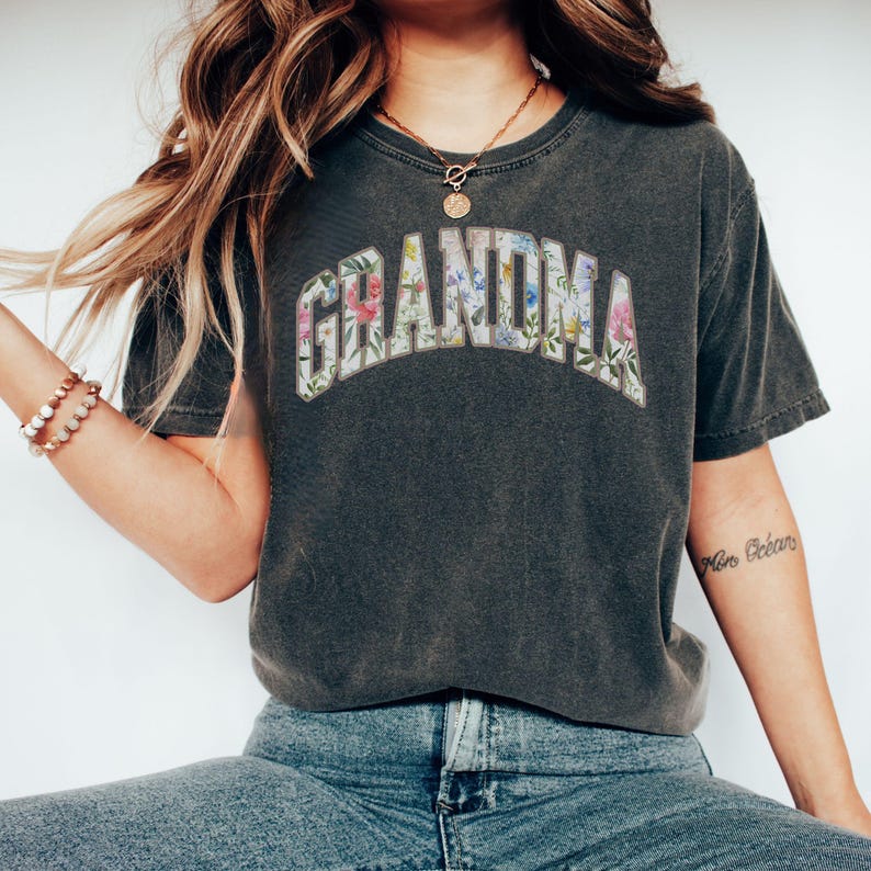 May include: A dark gray t-shirt with the word "Grandma" spelled out in white letters with a floral pattern. The flowers are pink, blue, and yellow.