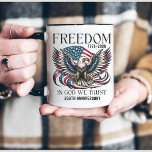 May include: White ceramic mug with a black handle and rim. The mug features the word "FREEDOM" above an illustration of an eagle with outstretched wings, the American flag, and the text "IN GOD WE TRUST 250TH ANNIVERSARY".