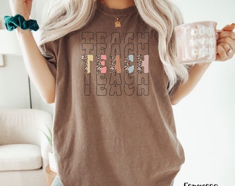 May include: A brown t-shirt with the word "TEACH" repeated four times in a stacked design. The word "TEACH" is outlined in black and filled with a leopard print pattern in pink, blue, and yellow.