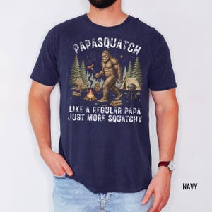 May include: Navy blue t-shirt featuring a Bigfoot graphic with a hot dog and campfire, and the text "PAPASQUATCH LIKE A REGULAR PAPA JUST MORE SQUATCHY."