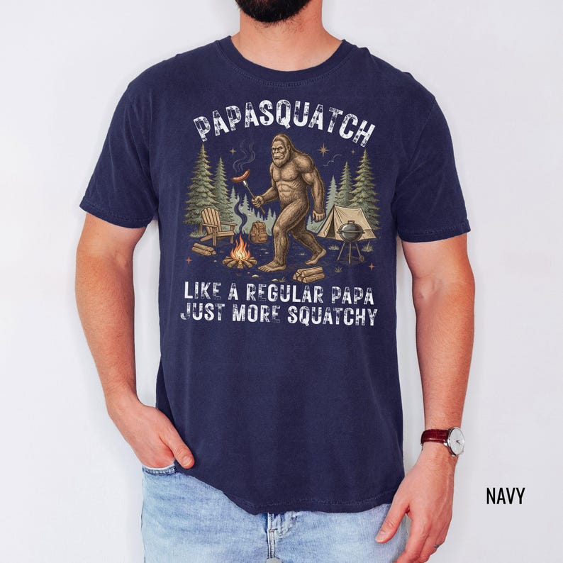May include: Navy blue t-shirt featuring a Bigfoot graphic with a hot dog and campfire, and the text "PAPASQUATCH LIKE A REGULAR PAPA JUST MORE SQUATCHY."