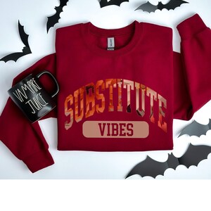 May include: A maroon sweatshirt with the text "SUBSTITUTE VIBES" in a large, orange, floral font. The sweatshirt is laid flat on a white surface with black bats scattered around it. A black mug with the text "VAMPIRE JUICE" is also on the surface.