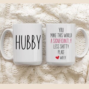 May include: Two white coffee mugs with black text. One mug says "Hubby" and the other mug says "You make this world a significantly less shitty place.  Wifey".