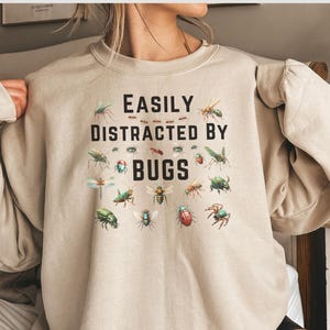 May include: A beige sweatshirt with the text "Easily Distracted by Bugs" printed on the front. The text is surrounded by a variety of colorful bug illustrations.