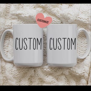 May include: Two white ceramic mugs with black text that reads "CUSTOM" on each mug. The mugs are on a white knitted surface.