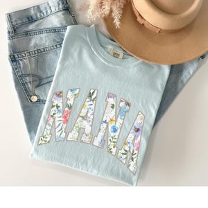 May include: A light blue t-shirt with the word "NANA" in a floral design. The shirt is paired with light wash jeans, a gold necklace, and a tan hat. A sprig of dried flowers is also visible.