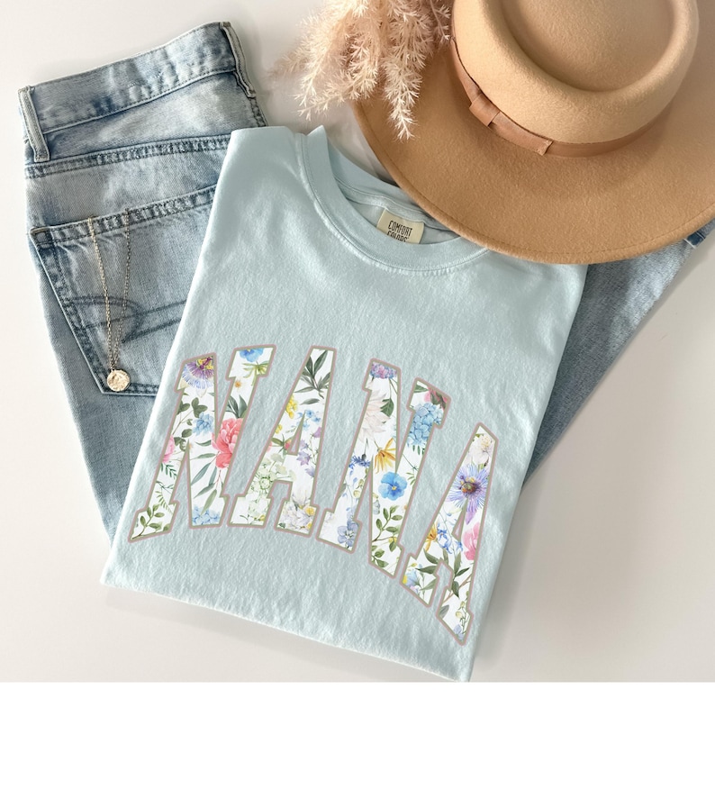 May include: A light blue t-shirt with the word "NANA" in a floral design. The shirt is paired with light wash jeans, a gold necklace, and a tan hat. A sprig of dried flowers is also visible.