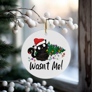 May include: White ceramic Christmas ornament with a black cat wearing a Santa hat tangled in Christmas lights and a Christmas tree. The ornament reads "Wasn't Me!"