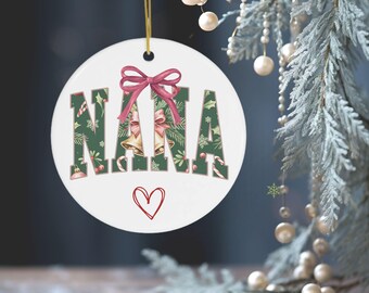 May include: A white, round Christmas ornament with the word "NANA" in green letters decorated with holly, candy canes, and bells. A red bow sits above the letters, and a red heart is drawn below. The ornament hangs from a gold string.
