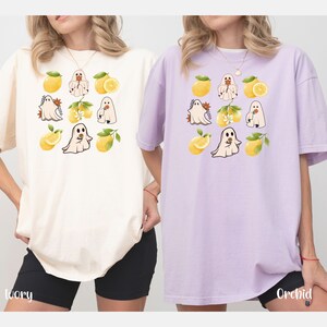 May include: Two women wearing oversized t-shirts with a lemon and ghost design. The shirt on the left is ivory and the shirt on the right is orchid.