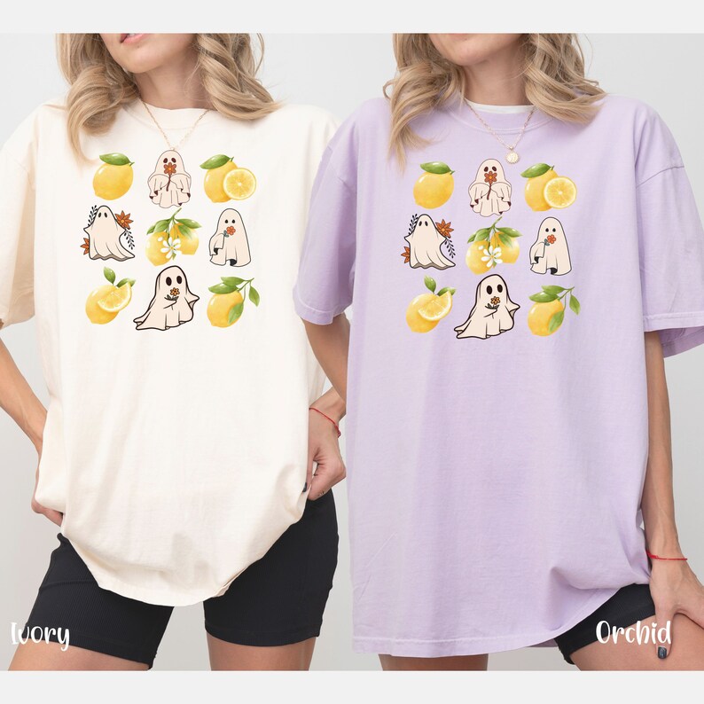 May include: Two women wearing oversized t-shirts with a lemon and ghost design. The shirt on the left is ivory and the shirt on the right is orchid.
