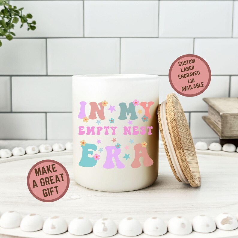 Op de afbeelding: Een cr&egrave;mekleurige keramische pot met een houten deksel, met de tekst "IN MY EMPTY NEST ERA" in kleurrijke letters in retrostijl. De pot heeft een ronde, cilindrische vorm en is versierd met bloemen en sterren. De afbeelding bevat ook twee ronde stickers met tekst.