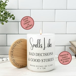 May include: A white candle with the text "Smells Like Bad Decisions & Good Stories" is lit, with a wooden lid beside it. The lid is engraved with "Custom Engraved Lid Available". The candle is printed on the candle, not stickers or vinyl.