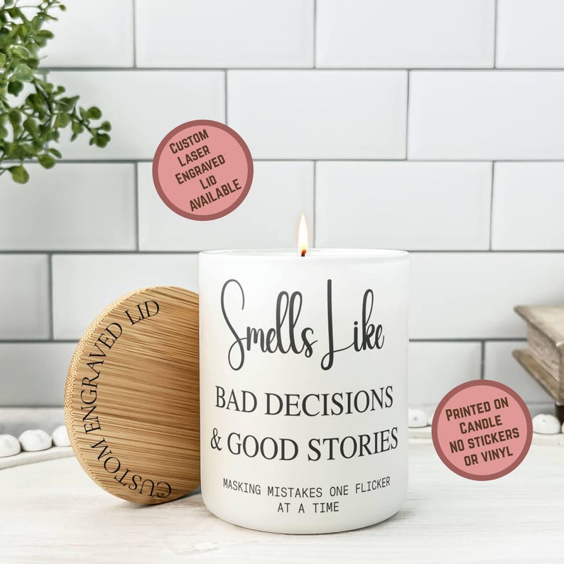 May include: A white candle with the text "Smells Like Bad Decisions & Good Stories" is lit, with a wooden lid beside it. The lid is engraved with "Custom Engraved Lid Available". The candle is printed on the candle, not stickers or vinyl.