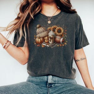 May include: A dark gray t-shirt with a graphic print of a fall-themed design. The design includes a pumpkin, a beanie, a coffee cup, a football, and sunflowers.