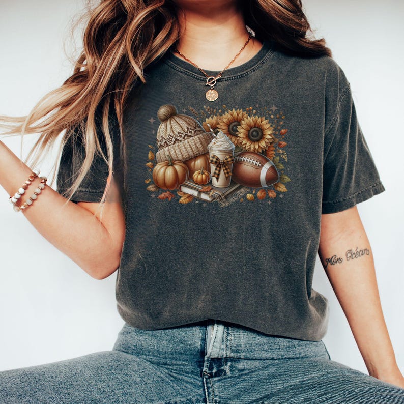May include: A dark gray t-shirt with a graphic print of a fall-themed design. The design includes a pumpkin, a beanie, a coffee cup, a football, and sunflowers.