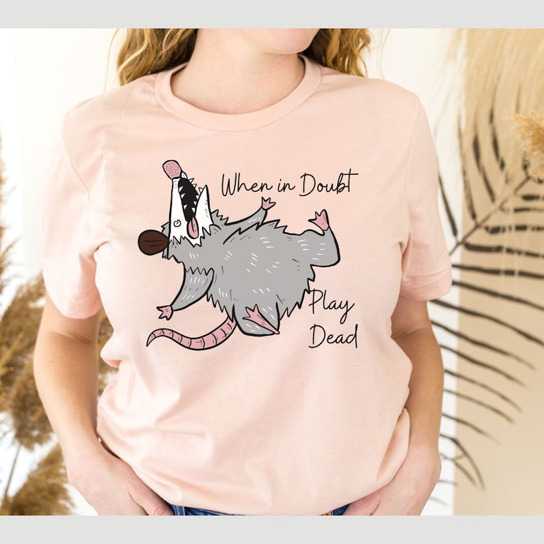 Pu&ograve; includere: Una maglietta rosa con un opossum dei cartoni animati che finge di essere morto. Il testo "When in doubt, play dead" &egrave; stampato sulla maglietta.