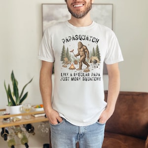 May include: White t-shirt featuring a Bigfoot graphic with a hot dog, and the text "Papasquatch Like a Regular Papa Just More Squatchy". The design includes a campfire, tent, and trees.