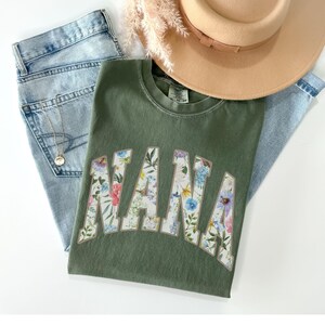 May include: An olive green t-shirt with the word "NANA" in a floral design, paired with light wash denim jeans and a tan hat. A gold necklace is visible on the jeans. The shirt is the focal point of the image.