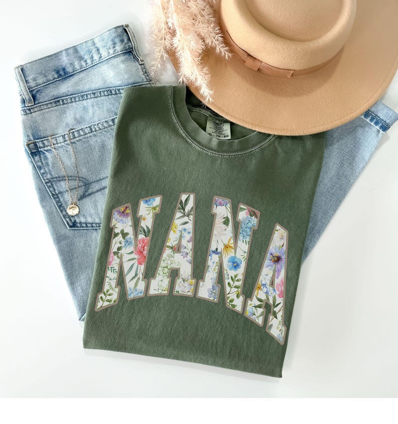 May include: An olive green t-shirt with the word "NANA" in a floral design, paired with light wash denim jeans and a tan hat. A gold necklace is visible on the jeans. The shirt is the focal point of the image.